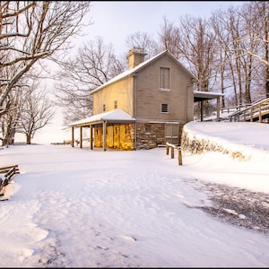 May include: A rustic, stone and wood building with a porch sits on a snowy hill. The building is surrounded by bare trees and a wooden fence. The snow is deep and white, and the sky is a pale blue.