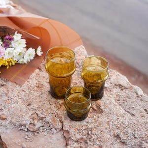 Peut inclure: Trois verres à boire ambrés de différentes tailles sont disposés sur une surface texturée brun clair. Les verres ont une forme cylindrique avec une bande décorative en haut et en bas. Un bouquet de fleurs dans du papier brun est en arrière-plan.