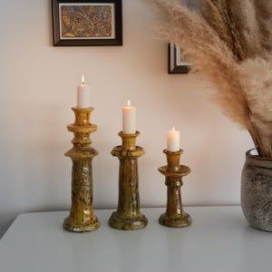 以下が含まれることがあります： 高さの異なる3つのセラミックキャンドルホルダー。それぞれに火の灯った白いキャンドルが立っています。ホルダーはまだらの黄褐色で、表面はざらざらしています。額入りのアートワークと、乾燥した植物が入った花瓶が背景にあります。