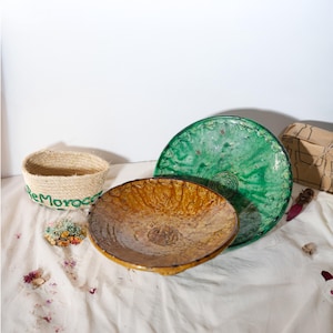 Op de afbeelding: Een verzameling handgemaakte aardewerk en geweven items. Bevat een groene keramische schaal, een geelbruine keramische kom en een kleine geweven mand met de tekst "reMorocco" in groen. De items zijn gerangschikt op een witte doek.