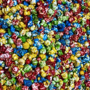 May include: A close-up of a large bowl filled with colorful popcorn. The popcorn is coated in a variety of colors, including red, blue, green, and yellow.