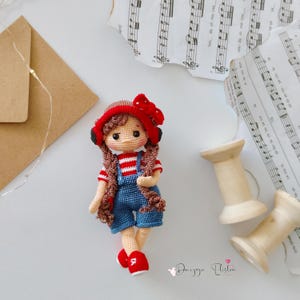 Puede incluir: Muñeca de ganchillo hecha a mano con trenzas marrones, que lleva un sombrero rojo con un lazo, una camisa a rayas rojas y blancas, un peto azul y zapatos rojos. La muñeca mide aproximadamente 15 cm.