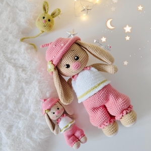 May include: Two crocheted bunny dolls, one pink and one white, wearing pink and white outfits. The larger bunny is wearing a pink hat and has long ears. The smaller bunny is wearing a pink hat and has short ears. Both bunnies have pink noses and black eyes.