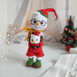 May include: A crocheted snowman doll wearing a red and green hat and holding a smaller crocheted snowman doll. The larger snowman has a yellow scarf and glasses.