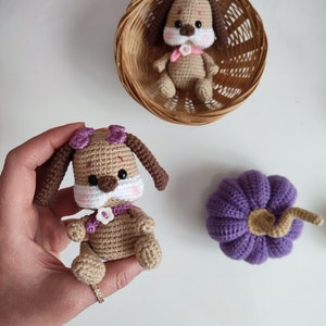 May include: Two crocheted brown and white dog plushies with purple bows. One dog is sitting in a wicker basket and the other is being held in a hand. A purple crocheted pumpkin is also in the image.