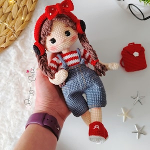 Puede incluir: Una muñeca de crochet con pelo rojo, un lazo rojo y una camisa a rayas rojas y blancas. La muñeca lleva un mono azul y zapatos rojos. La muñeca lleva una pequeña bolsa roja con una flor blanca.