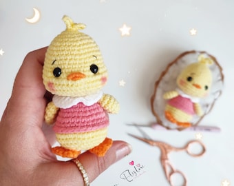 May include: A crocheted yellow chick wearing a pink dress with a white collar. The chick is holding a small pink heart in its hand. The chick is sitting on a white surface.