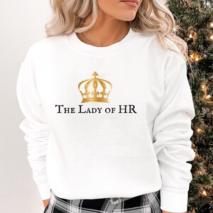 Peut inclure: Sweat-shirt blanc avec une couronne dorée et le texte "The Lady of HR" imprimé sur le devant.