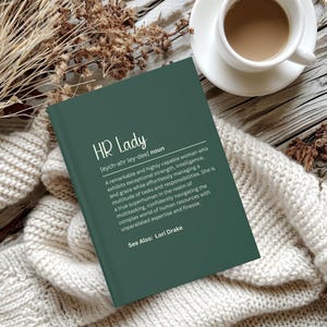 Op de afbeelding: Een groen boek met de titel "HR Lady" en een definitie van de term. Het boek ligt op een wit gebreid oppervlak.