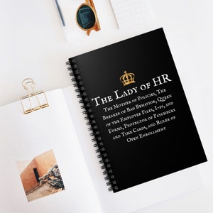 Può includere: Un taccuino nero a spirale con una corona dorata e il testo "The Lady of HR" in bianco. Il testo sotto il titolo recita: "The Mother of Policies, The Breaker of Bad Behavior, Queen of the Employee Files, I-95, and Forms, Protector of Paychecks and Time Cards, and Ruler of Open Enrollment."
