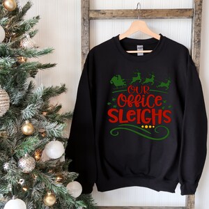 Peut inclure: Sweat-shirt noir avec un motif vert et rouge qui dit "Our Office Sleighs" avec un graphique de Père Noël et de rennes.