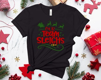 Może przedstawiać: Czarny T-shirt z zielonym i czerwonym wzorem przedstawiającym świąteczną scenę z reniferami i tekstem "Our Team Sleighes".