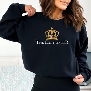 Peut inclure: Sweat-shirt noir avec une couronne dorée et le texte "The Lady of HR" imprimé sur le devant.