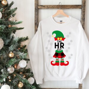 Peut inclure: Sweat-shirt blanc de Noël avec un motif d'elfe en plaid rouge et noir. L'elfe porte un chapeau vert et a les mots "The HR Elf" écrits sur sa chemise.