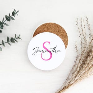 Könnte beinhalten: Runder weißer Untersetzer mit einem rosa Buchstaben "S" und dem Namen "Samantha" in schwarzer Schrift. Der Untersetzer liegt auf einer weißen Oberfläche.