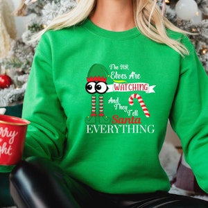 Peut inclure: Sweat-shirt de Noël vert avec un lutin de dessin animé portant un chapeau vert et tenant une canne en sucre. Le texte sur le sweat-shirt est "The HR Elves Are Watching And They Tell Santa EVERYTHING."