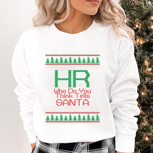 Peut inclure: Sweat-shirt blanc avec un motif de Noël vert et rouge. Le motif représente un "HR" stylisé avec le texte "Who Do You Think Tells Santa".