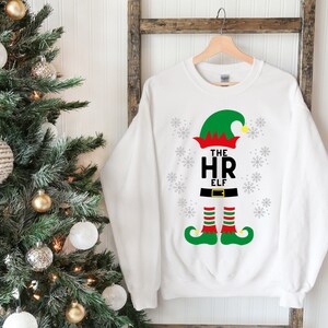 Peut inclure: Pull-over blanc de Noël avec un motif d'elfe vert. L'elfe porte un chapeau rouge avec un pompon blanc, un costume vert et des bas rayés rouge et blanc. Le texte "The HR Elf" est imprimé sur le pull.
