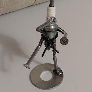 May include: A small metal sculpture of a person made from various metal parts, including a spark plug, screws, and washers. The sculpture is standing on a round metal base.