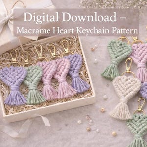 以下が含まれることがあります： パステルカラーのラベンダー、ピンク、ミントグリーンのマクラメハートキーホルダー。ゴールドカラーの留め金とタッセル付き。キーホルダーはギフトボックスに入れられ、明るい色の表面に配置されています。「Digital Download - Macrame Heart Keychain Pattern」と書かれたテキスト。