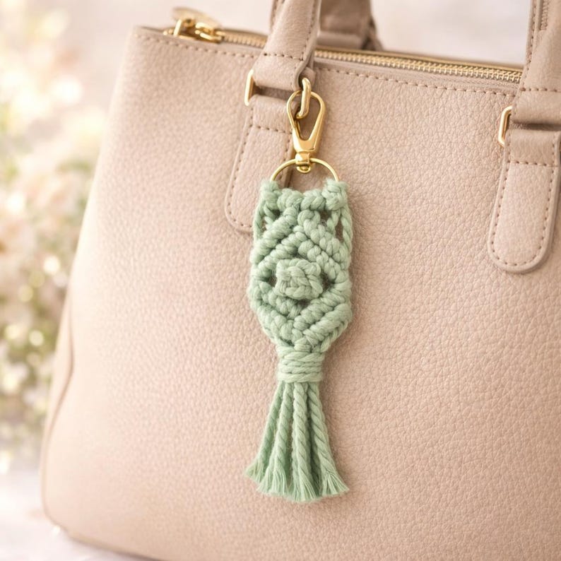 Pu&ograve; includere: Un portachiavi in macram&egrave; verde chiaro con chiusura color oro, attaccato a una borsa in pelle beige. Il portachiavi presenta un design intrecciato con una nappa nella parte inferiore. La borsa ha una chiusura a zip e un manico.