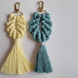 May include: Two macrame keychains featuring knotted cord designs in pale yellow and muted teal. Each keychain includes a gold-toned metal lobster clasp attached to a circular ring, with a long, flowing tassel extending from the base of the knots.