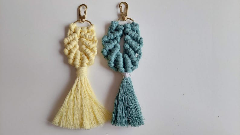 May include: Two macrame keychains featuring knotted cord designs in pale yellow and muted teal. Each keychain includes a gold-toned metal lobster clasp attached to a circular ring, with a long, flowing tassel extending from the base of the knots.