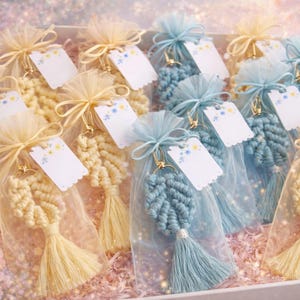 May include: A collection of macrame leaf keychains in cream and blue, each individually packaged in a sheer organza drawstring bag with a small white gift tag featuring a floral print, all arranged together in a rectangular box.