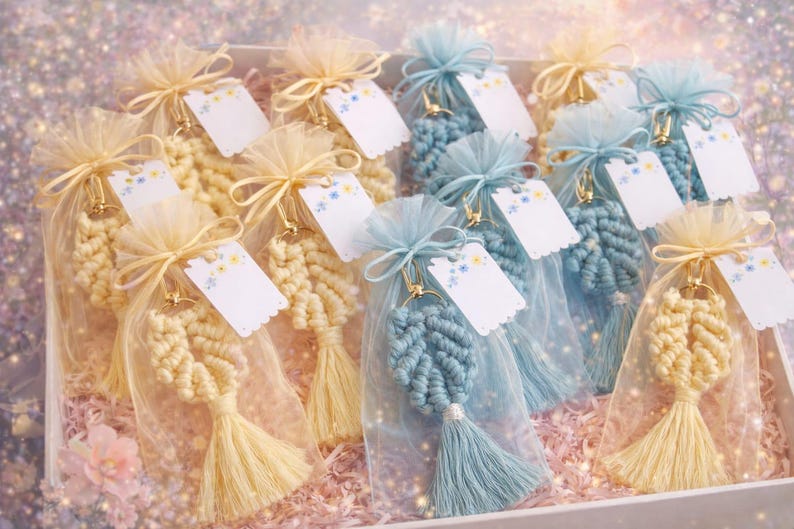 May include: A collection of macrame leaf keychains in cream and blue, each individually packaged in a sheer organza drawstring bag with a small white gift tag featuring a floral print, all arranged together in a rectangular box.