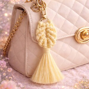 May include: A cream-coloured macrame bag charm featuring a knotted design and a long tassel, attached to a gold-tone metal lobster clasp. The charm hangs from the gold-tone chain and hardware of a quilted, light beige handbag with a gold-tone clasp.