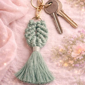 May include: A sage green macrame keyring featuring a knotted leaf-shaped design attached to a gold-toned metal lobster clasp and split ring. Two silver-toned metal keys are attached to the ring, and a long, straight tassel hangs from the bottom.
