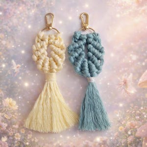 May include: Two macrame keychains featuring knotted cord designs and long tassels. One keychain is cream-coloured, and the other is dusty blue. Both are attached to gold-toned metal lobster claw clasps and circular rings for secure attachment.