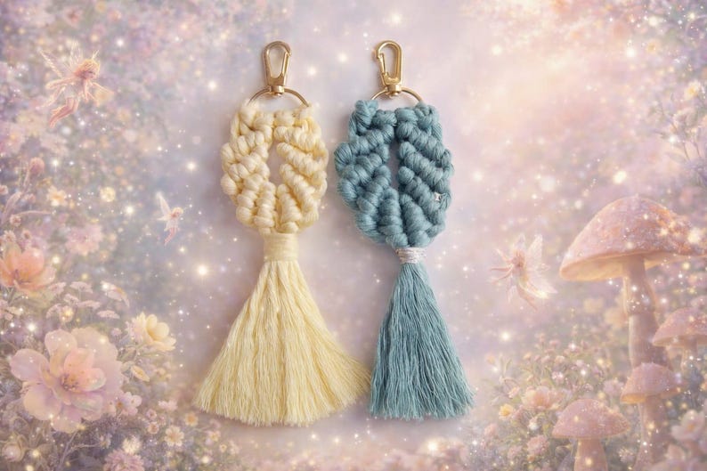 May include: Two macrame keychains featuring knotted cord designs and long tassels. One keychain is cream-coloured, and the other is dusty blue. Both are attached to gold-toned metal lobster claw clasps and circular rings for secure attachment.