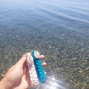 Op de afbeelding: Twee macramé sleutelhangers, één witte en één turquoise, elk versierd met een zeeschelp. De sleutelhangers worden omhoog gehouden tegen een achtergrond van een heldere, zonnige zee met een boot in de verte.