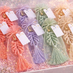 May include: A collection of macrame leaf keychains in coral, lavender, sage green, and cream, each encased in a matching sheer organza drawstring bag. Every keychain features a gold-tone metal clasp and a small white scalloped gift tag.