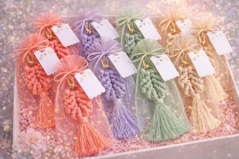 May include: A collection of macrame leaf keychains in coral, lavender, sage green, and cream, each encased in a matching sheer organza drawstring bag. Every keychain features a gold-tone metal clasp and a small white scalloped gift tag.
