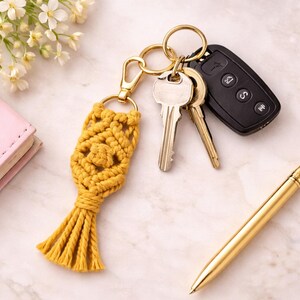 Pu&ograve; includere: Portachiavi in macram&egrave; giallo senape con nappa. Il portachiavi &egrave; attaccato a un moschettone e un portachiavi color oro, che tiene chiavi e un telecomando per auto. Una penna dorata e un piccolo taccuino rosa sono anch'essi nell'immagine.