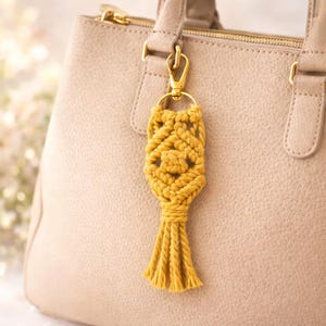 Pu&ograve; includere: Portachiavi in macram&egrave; giallo senape con chiusura color oro, attaccato a una borsa beige. Il portachiavi presenta un design intrecciato con una nappa in basso. La borsa ha una superficie strutturata.