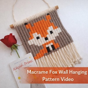 以下が含まれることがあります： オレンジ、白、黒、茶色でピクセル化されたデザインの、マクラメのキツネの壁掛け。壁掛けの上部には木の棒、下部にはフリンジが付いています。「Macrame Fox Wall Hanging Pattern Video」の文字が見えます。
