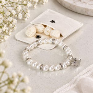 May include: A white pearl bracelet featuring silver-toned spacers embedded with small clear crystals. The bracelet includes a silver-toned metal clasp and is positioned in front of a small, white, snap-button fabric pouch containing four white stones.