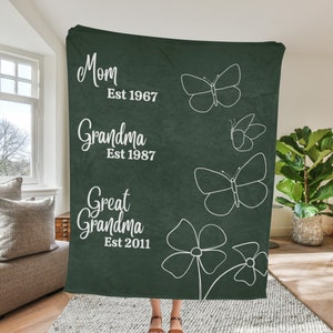 May include: A dark green fleece blanket with white text and butterfly designs. The text reads "Mom Est 1967, Grandma Est 1987, Great Grandma Est 2011".