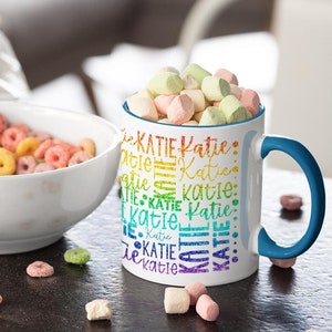 Pode incluir: Uma caneca de cerâmica branca com uma alça azul. A caneca tem um design de arco-íris com o nome "Katie" repetido várias vezes. A caneca está cheia de marshmallows de cores pastel e está sobre uma superfície marrom escura com anéis de cereal espalhados.