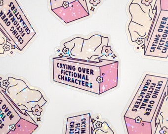 May include: A set of six pink and white holographic stickers with the text "Crying Over Fictional Characters" and a cartoon image of a tissue box.