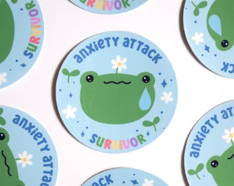 May include: A set of six round stickers featuring a cartoon frog with a flower on its head and a tear on its cheek. The stickers have a blue background with the text "anxiety attack survivor" in rainbow colors.