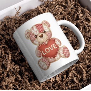 以下が含まれることがあります： 「LOVE」の文字が入った赤いハートを持った、パッチワークのテディベアが描かれた白いセラミックマグ。クマはピンクと赤のチェック柄と花柄です。マグカップは、茶色の紙の詰め物が入った白い箱の中に入っています。