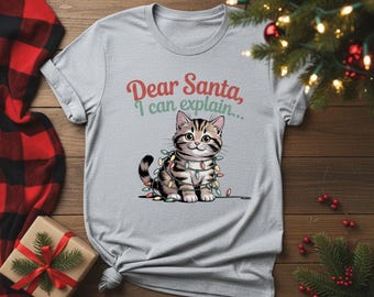 May include: A light gray t-shirt featuring a cartoon kitten adorned with Christmas lights. The text "Dear Santa, I can explain..." is printed above the cat. The shirt is displayed on a wooden surface with a plaid blanket and a wrapped gift.
