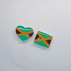 May include: Two decorative pins featuring the Jamaican flag. One pin is heart-shaped, and the other is a square with a diagonal design. The pins have a green, black, and orange color scheme.