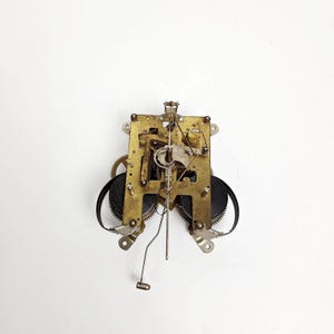 May include: A vintage clock movement with brass gears, black weights, and a pendulum. The mechanical components are arranged on a rectangular brass plate with multiple mounting holes. The clock mechanism is set against a white background.