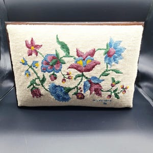 May include: A rectangular needlepoint pillow featuring a cream-colored background with a floral pattern in shades of blue, pink, green, and yellow. The design includes various stylized blossoms and leaves, with the signature M. LERNER 86 in the corner.