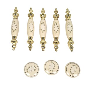 May include: A set of five elongated cabinet pulls and three circular cabinet knobs. Each piece features a cream-colored ceramic center with a delicate green and white floral pattern, accented by polished gold-tone metal hardware at the ends.
